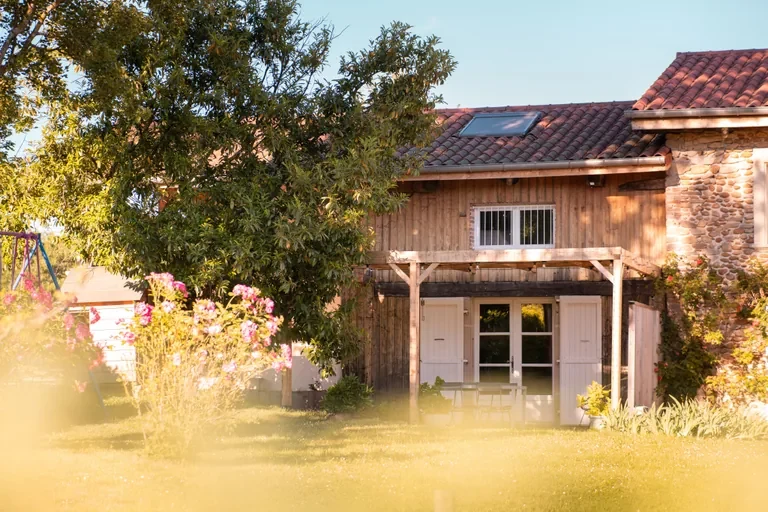 Façade en pierre du gîte L'Hospitalier avec roses, pergola et jardin en lumière de fin de journée