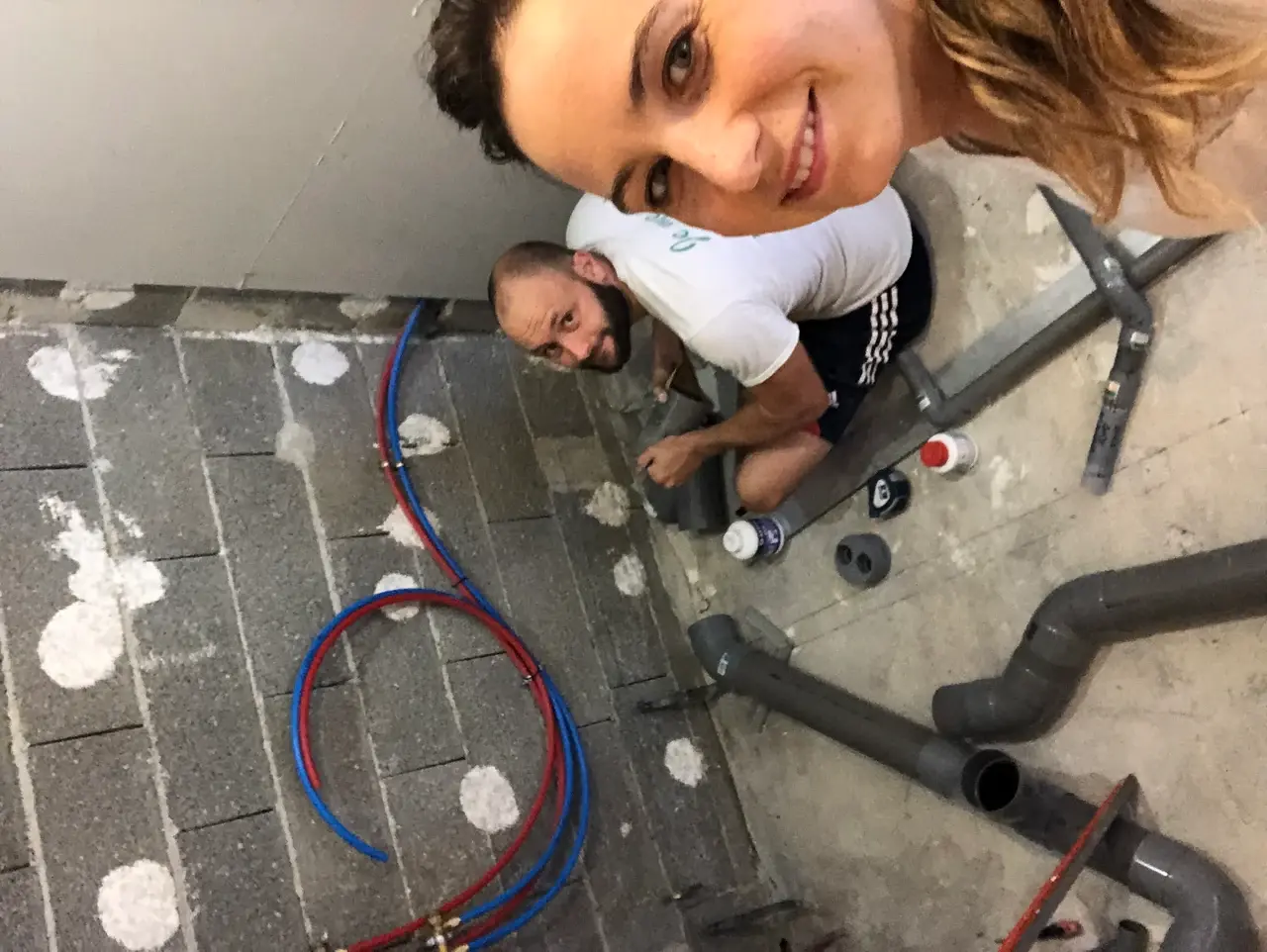 Esther souriante en selfie, Arnaud en train de poser la plomberie derrière elle sur le chantier