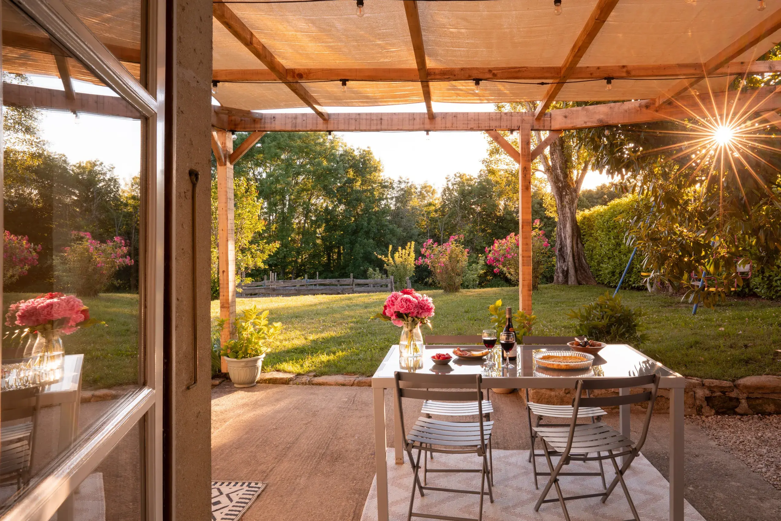 Vue depuis l'interieur du gite sur la terrasse, la table dressee et le jardin avec soleil en etoile